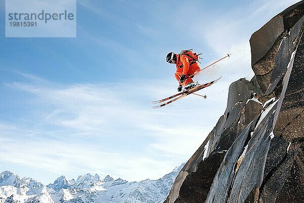 Ein professioneller Skifahrer macht einen Absprung von einer hohen Klippe gegen den blaün Himmel und hinterlässt eine Spur von Pulverschnee in den Bergen. Kaukasischer Bergrücken im Hintergrund. Foto von den Hängen des Berges Elbrus. Das Konzept des Extremsports und der Freizeitgestaltung in den