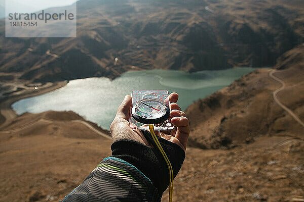 Ein Mann hält einen Taschenmagnetkompass zur Navigation vor dem Hintergrund eines felsigen Abhangs und eines Bergsees in der Hand. Das Konzept  einen Weg zu finden. Freiheit gewinnen. Freiheit der Wahl