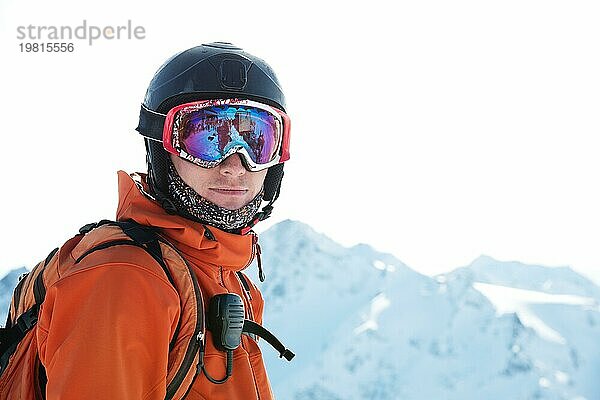 Porträt eines Skifahrers in einem orangefarbenen Overall mit einem Rucksack auf dem Rücken in einem Helm steht vor dem Hintergrund einer schönen kaukasischen Berglandschaft mit schneebedeckten Bergen des kaukasischen Kammes und blaün Himmel. Kopieren Sie das Konzept der aktiven recreati