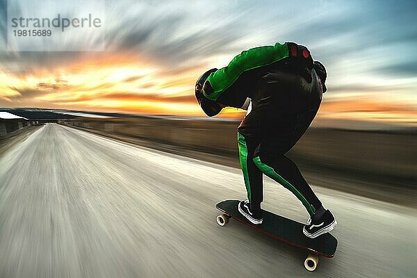 Ein Mann in Helm und Lederkombi fährt in einem speziellen Gestell mit hoher Geschwindigkeit auf einem Longboard bergab auf Afsalt vor der Kulisse eines schönen Sonnenuntergangshimmels im Gegenlicht der untergehenden Sonne. Der Hintergrund ist dynamisch aufgefächert