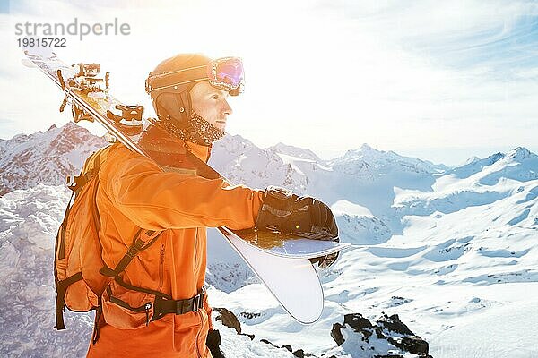 Porträt eines Skifahrers in einem orangefarbenen Overall mit einem Rucksack auf dem Rücken und Skiern auf seinen Schultern in einem Helm steht auf einem Felsen gegen den blaün Himmel. Kopieren Sie den Raum. Das Konzept der aktiven Erholung im Winter und Skifahren