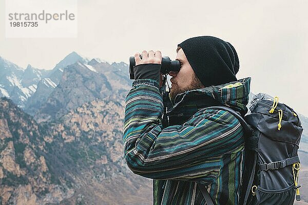 Ein Hipster Mann mit Bart  Hut  Jacke und Rucksack in den kaukasischen Bergen hält ein Fernglas  Abenteuer  Tourismus  Fährtensuche