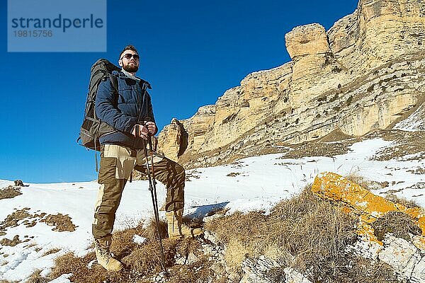 Ein Hipster Reisender mit Bart und Sonnenbrille in der Natur. Ein Mann wandert in den Bergen mit einem Rucksack und skandinavischen Wanderstöcken vor dem Hintergrund einer Berglandschaft und blauem Himmel. Reisen Lifestyle Abenteuer Outdoor Erholung Outdoor Sport