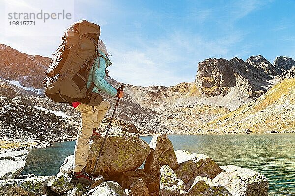 Schlankes und sympathisches Wandermädchen mit Sonnenbrille und blaür dünner Daunenjacke mit Rucksack und Trekkingstöcken steigt auf einen hohen Felsen vor dem Hintergrund von Felsen und einem hohen Bergsee