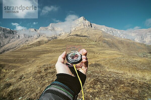 Blickpunktaufnahme. Eine Ichperspektive eines Mannes  der einen Kompass vor dem Hintergrund einer epischen Landschaft mit Klippen  Hügeln und einem blaün Himmel mit Wolken hält