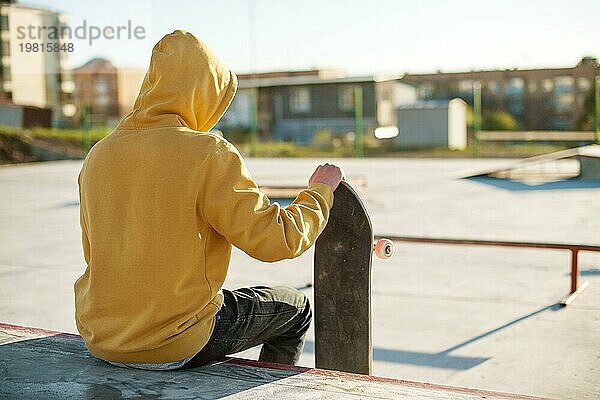 Nahaufnahme eines jungen Hipsters in Sweatshirt und Jeans  der traurig in einem Skatepark sitzt und ein Skateboard hält