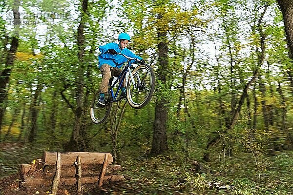 Ein junger Fahrer mit Helm und blauem Sweatshirt fliegt auf einem Fahrrad  nachdem er von einem hohen Kicker auf einem Waldradweg gesprungen ist. Aufnahme mit Langzeitbelichtung Verdrahtung