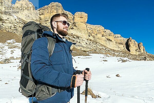 Ein Hipster Reisender mit Bart und Sonnenbrille in der Natur. Ein Mann wandert in den Bergen mit einem Rucksack und skandinavischen Wanderstöcken vor dem Hintergrund einer Berglandschaft und blauem Himmel. Reisen Lifestyle Abenteuer Outdoor Erholung Outdoor Sport