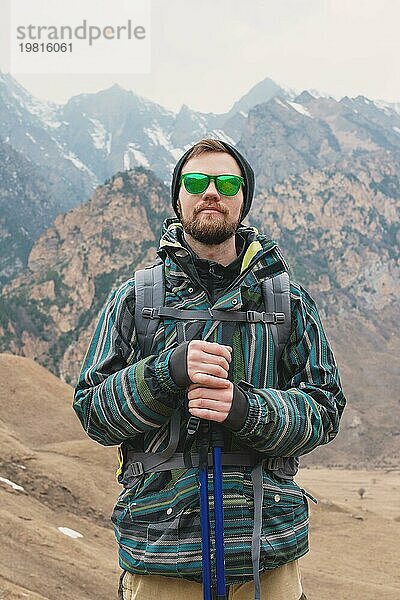 Ein Mann mit Bart und Sonnenbrille in einer Membranjacke  Hut  mit einem Rucksack und Stöcken für Nordic Walking  ein Reisender  der in der freien Natur steht und auf die Berge schaut