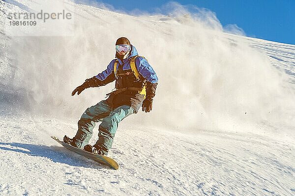 Ein Freeboard Snowboarder mit Skimaske und Rucksack fährt über die schneebedeckte Piste und hinterlässt einen Pulverschnee vor dem blaün Himmel