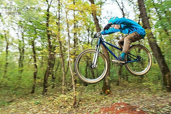 Ein junger Fahrer mit Helm und blauem Sweatshirt fliegt auf einem Fahrrad  nachdem er von einem hohen Kicker auf einem Waldradweg gesprungen ist. Aufnahme mit Langzeitbelichtung Verdrahtung