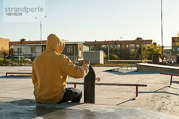 Nahaufnahme eines jungen Hipsters in Sweatshirt und Jeans  der traurig in einem Skatepark sitzt und ein Skateboard hält