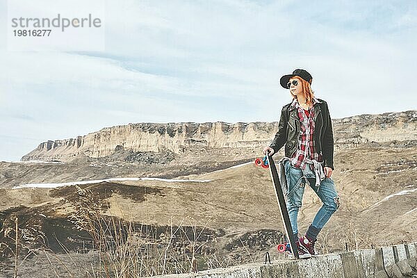 Stylische rothaarige Frauen in Jeansoveralls stehen mit einem Longboard auf einer Bodenwelle am Highway in den Bergen