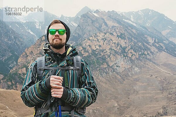 Ein Mann mit Bart und Sonnenbrille in einer Membranjacke  Hut  mit einem Rucksack und Stöcken für Nordic Walking  ein Reisender  der in der freien Natur steht und auf die Berge schaut