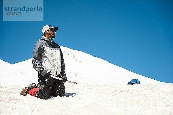 Training zum Korrigieren des Ausrutschens an einem Hang oder auf einem Gletscher mit Hilfe eines Eispickels. Ein voll ausgerüsteter Rucksacktourist kniet auf einem verschneiten Hang in den Bergen in der Nähe des Eispickels. Im Hintergrund beobachten Bergsteiger  die einen unerfahrenen Kletterer vor dem Aufstieg trainieren