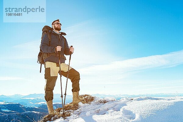 Ein Hipster Reisender mit Bart und Sonnenbrille in der Natur. Ein Mann wandert in den Bergen mit einem Rucksack und skandinavischen Wanderstöcken vor dem Hintergrund einer Berglandschaft und blauem Himmel. Reisen Lifestyle Abenteuer Outdoor Erholung Outdoor Sport