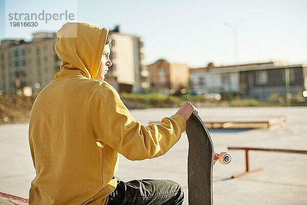 Nahaufnahme eines jungen Hipsters in Sweatshirt und Jeans  der traurig in einem Skatepark sitzt und ein Skateboard hält