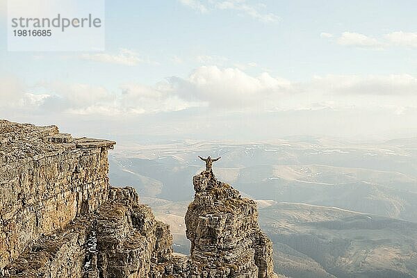 Ein glücklicher Mann mit hoch erhobenen Händen steht auf einem separat stehenden Felsen  der sich über den Wolken befindet  vor dem Hintergrund von Tälern  Hügeln  Sonnenuntergang und einem schönen Himmel