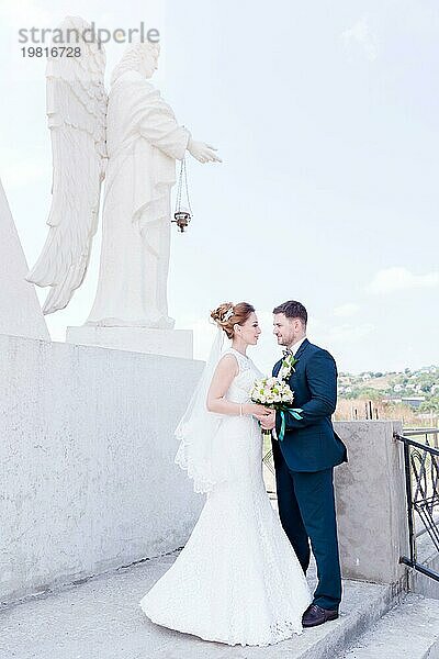 Porträt eines schönen Paares in den Flitterwochen an einem Hochzeitstag mit einem Blumenstrauß in der Hand vor dem Hintergrund eines orthodoxen christlichen Denkmal mit Engeln. Das Konzept der christlichen Hochzeit in der Kirche der Frischvermählten und der spirituelle Glaube der jungen Familie