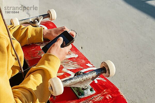 Nahaufnahme eines Teenagers in Sweatshirt  Jeans und Turnschuhen  der in einem Skatepark sitzt und ein Telefon mit Kopfhörern und ein Skateboard hält