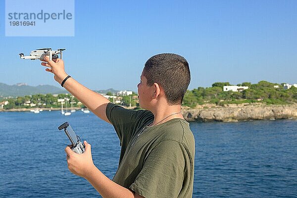 Glücklicher Teenager  der die Flugdetails für den Drohnenflug vorbereitet und abschließt  während eines sonnigen Tages mit dem Mittelmeer im Hintergrund Spanien  Balearen