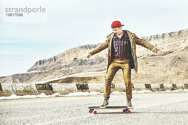 Stylish happy Junger Mann in Mütze und Hose Jogger rollen auf einem Longboard eine Bergstraße hinunter  genießen das Leben