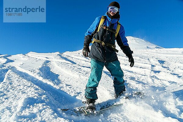 Ein Freeboard Snowboarder mit Skimaske und Rucksack fährt über die schneebedeckte Piste und hinterlässt einen Pulverschnee vor dem blaün Himmel