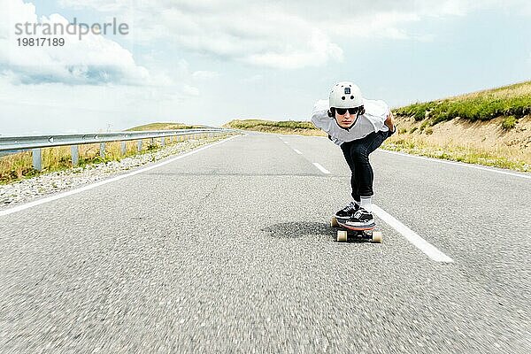 Der Typ mit dem Helm und der Sonnenbrille fährt mit seinem Longboard in einem speziellen Sportständer sehr schnell über die asphaltierte Straße