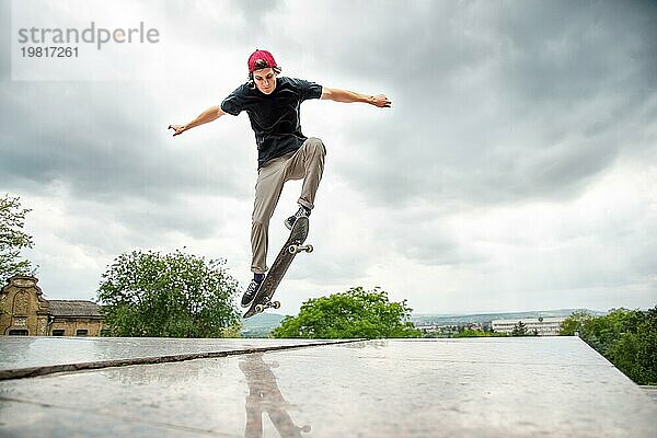 Langhaariger Skater Teenager in TShirt und Turnschuhmütze springt den Ollie vor dem Hintergrund eines stürmischen Himmels über der Stadt