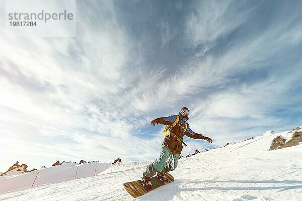 Ein Snowboarder mit Skimaske und Rucksack fährt auf einer schneebedeckten Piste und hinterlässt einen Pulverschnee vor dem blaün Himmel und der untergehenden Sonne. Foto in Bewegung