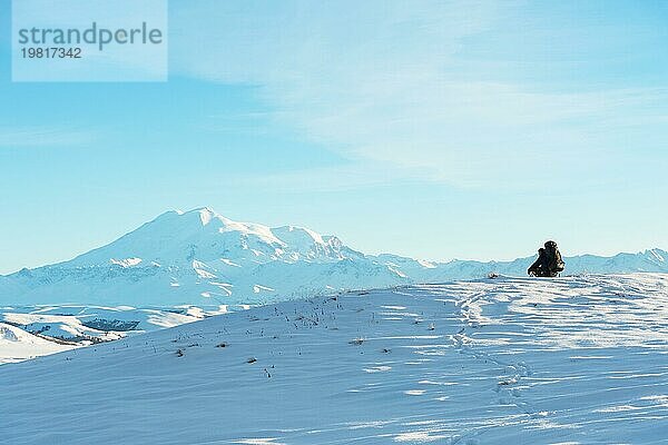 Ein Wanderer mit einem großen Rucksack auf den Schultern sitzt auf einem schneebedeckten Hügel vor dem blaün Himmel und dem schlafenden Vulkan Elbrus  der vom kaukasischen Hauptkamm umgeben ist  und meditiert. Das Konzept des Wanderns und der Harmonie durch Meditation in der Natur im Winter