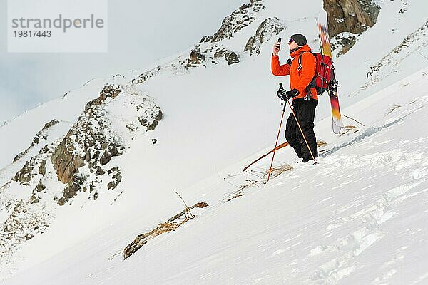 Ein Skifreerider  der auf einer Piste im Tiefschnee steht  fotografiert die Landschaft mit seinem Mobiltelefon  während er seine Ausrüstung auf dem Rücken trägt  die an einem Rucksack befestigt ist. Das Konzept des Winter Extremsports