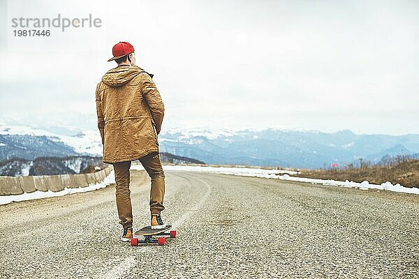 Stylish happy Junger Mann in Mütze und Hose Jogger rollen auf einem Longboard eine Bergstraße hinunter  genießen das Leben