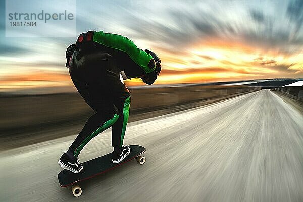 Ein Mann in Helm und Lederkombi fährt in einem speziellen Gestell mit hoher Geschwindigkeit auf einem Longboard bergab auf Afsalt vor der Kulisse eines schönen Sonnenuntergangshimmels im Gegenlicht der untergehenden Sonne. Der Hintergrund ist dynamisch aufgefächert