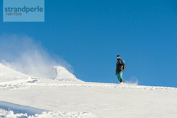 Ein Freeboard Snowboarder mit Skimaske und Rucksack fährt über die schneebedeckte Piste und hinterlässt einen Pulverschnee vor dem blaün Himmel