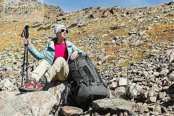 Ein Touristenmädchen mit Sonnenbrille  Daunenjacke und Hut  mit einem Rucksack und einer Bergausrüstung mit Griffen zum Wandern in den Händen  sitzt und entspannt sich und genießt die schöne Aussicht auf einen Hochgebirgssee im Nordkaukasus. Karatschai Tscherkess R