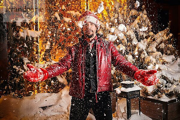 Mann gekleidet als Weihnachtsmann wirft Schnee auf einer Straße mit Girlanden geschmückt. Konzept für Neujahr und Weihnachten. Gemischte Medien