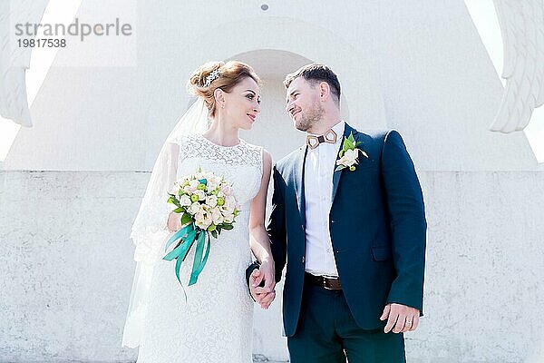 Porträt eines schönen Paares in den Flitterwochen an einem Hochzeitstag mit einem Blumenstrauß in der Hand vor dem Hintergrund eines orthodoxen christlichen Denkmal mit Engeln. Das Konzept der christlichen Hochzeit in der Kirche der Frischvermählten und der spirituelle Glaube der jungen Familie