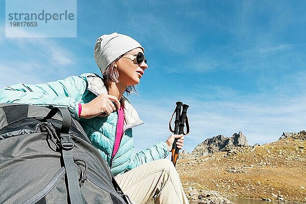 Ein Touristenmädchen mit Sonnenbrille  Daunenjacke und Hut  mit einem Rucksack und einer Bergausrüstung mit Griffen zum Wandern in den Händen  sitzt und entspannt sich und genießt die schöne Aussicht auf einen Hochgebirgssee im Nordkaukasus. Karatschai Tscherkess R