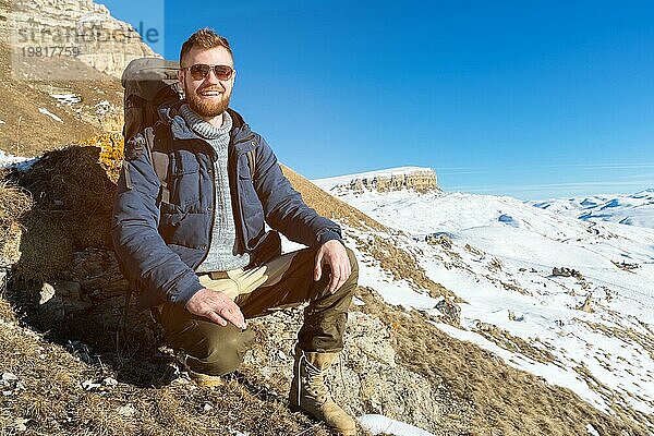 Porträt eines fröhlich lachenden Hipster Reisenden mit Bart und Sonnenbrille  der in der Natur sitzt. Ein Mann wandert in den Bergen mit einem Rucksack und skandinavischen Wanderstöcken im Hintergrund einer Berglandschaft. Reisen Lifestyle Abenteuer im Freien erholen