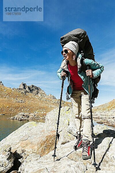 Schlankes und sympathisches Wandermädchen mit Sonnenbrille und blaür dünner Daunenjacke mit Rucksack und Trekkingstöcken steigt auf einen hohen Felsen vor dem Hintergrund von Felsen und einem hohen Bergsee