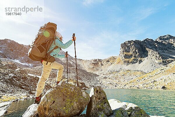 Schlankes und sympathisches Wandermädchen mit Sonnenbrille und blaür dünner Daunenjacke mit Rucksack und Trekkingstöcken steigt auf einen hohen Felsen vor dem Hintergrund von Felsen und einem hohen Bergsee