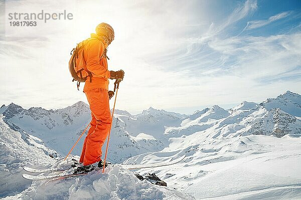 Ein Skifahrer in einem orangefarbenen Overall  mit einem Rucksack auf dem Rücken  einem Helm und Skistöcken in den Händen steht an einem Abgrund inmitten einer wunderschönen kaukasischen Berglandschaft mit schneebedeckten Gipfeln der Berge des Ca