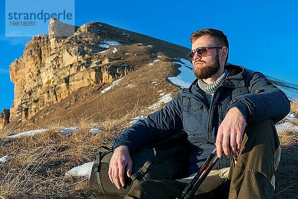 Porträt eines ruhigen Hipster Reisenden mit Bart und Sonnenbrille  der in der Natur sitzt. Ein Mann wandert in den Bergen mit einem Rucksack und skandinavischen Wanderstöcken im Hintergrund eines großen epischen Felsens. Reisen Lifestyle Abenteuer im Freien Erholung im Freien