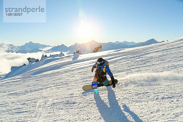 Ein Snowboarder mit Skimaske und Rucksack fährt auf einer schneebedeckten Piste und hinterlässt einen Pulverschnee vor dem blaün Himmel und der untergehenden Sonne. Foto in Bewegung