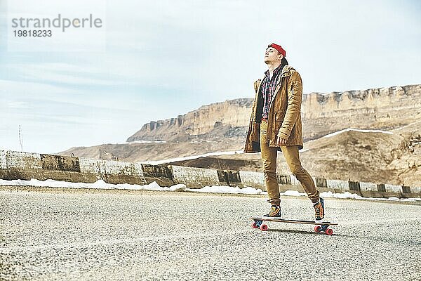 Stylish happy Junger Mann in Mütze und Hose Jogger rollen auf einem Longboard eine Bergstraße hinunter  genießen das Leben