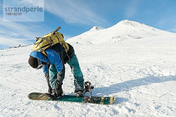 Ein Snowboarder mit einem Rucksack auf dem Rücken befestigt die Snowboardbindungen vor dem Hintergrund des schlafenden kaukasischen Vulkans Elbrus. Nordkaukasus