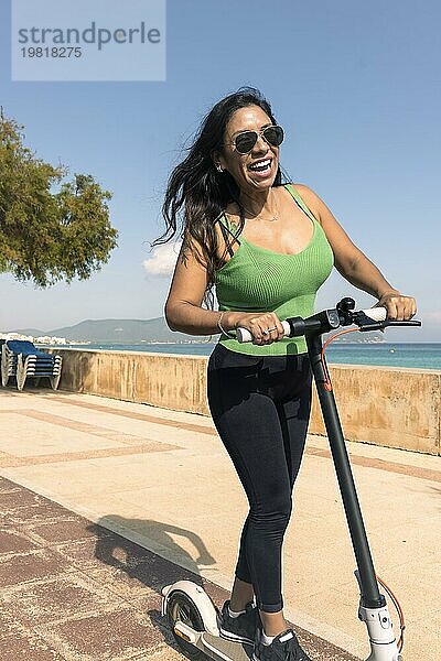 Junge Latina hat Spaß mit einem Elektroroller auf der Strandpromenade