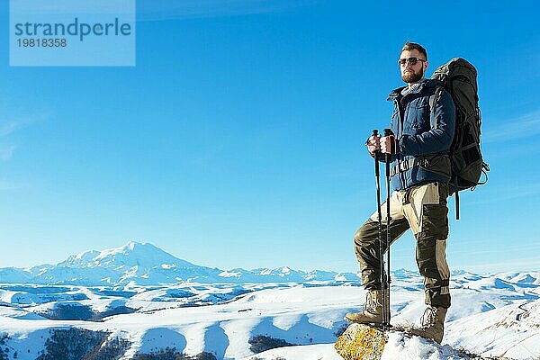 Ein Hipster Reisender mit Bart und Sonnenbrille in der Natur. Ein Mann wandern in den Bergen mit einem Rucksack und skandinavischen Wanderstöcke im Hintergrund eines Berges Elbrus Landschaft und blaün Himmel. Reisen Lifestyle Abenteuer Outdoor Erholung übertreffen