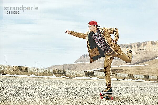 Stylish happy Junger Mann in Mütze und Hose Jogger rollen auf einem Longboard eine Bergstraße hinunter  genießen das Leben